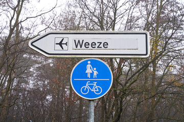 Street sign shows the way to the airport in Weeze. Bicycle lane and sidewalk sign hangs underneath.