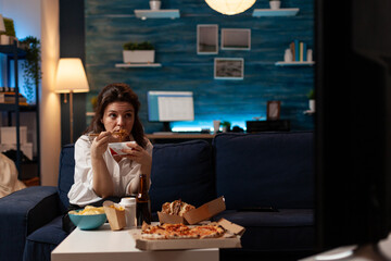 Woman sitting on couch eating takeout asian noodles with chopsticks watching television in home livingroom. Caucasian girl enjoying chinese takeaway ramen box and fast food delivery.