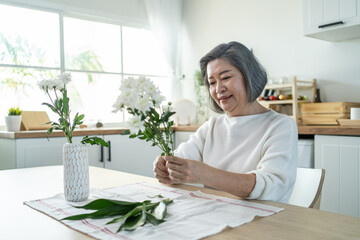Asian senior old woman puts flowers on vase with happiness in kitchen.