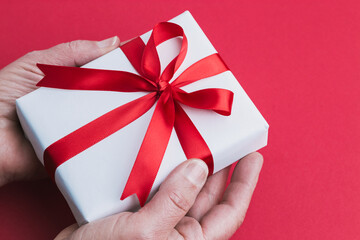 A two hands holding a small white gift with a red ribbon on a red background