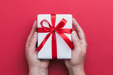 A two hands holding a small white gift with a red ribbon on a red background