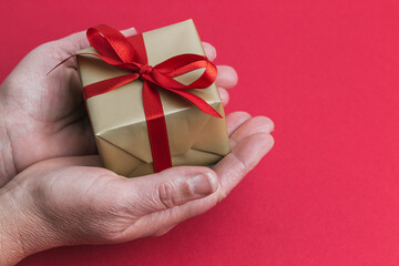 A two hands holding a small gold gift with a red ribbon on a red background