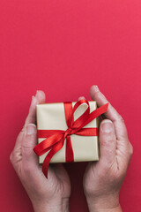 A two hands holding a small gold gift with a red ribbon on a red background