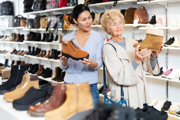 Female sales consultant helps a mature european woman choose winter shoes in a store