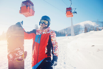 Happy young bearded man with helmet holding snowboard against backdrop of ski resort and lift...