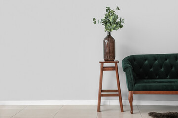 Vase with eucalyptus on wooden stool and green sofa near light wall