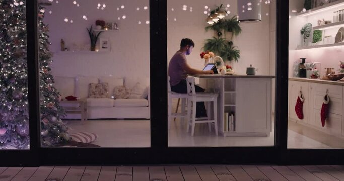 adult man working at the desk with a laptop at winter holidays and snow falling behind the glass sliding doors, cozy decorated apartment on Christmas