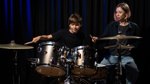 Young Caucasian Woman Teaches A Boy To Play The Drums In The Studio On A Black Background. Music School Student