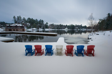 Colourful Adirondack chairs facing winter landscape covered in snow with a lake