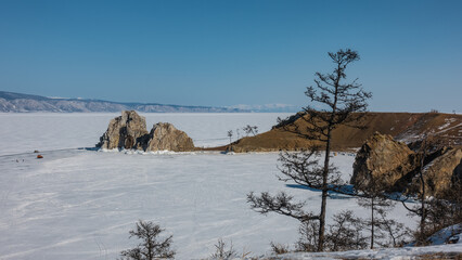 A two-headed rock, devoid of vegetation, rises above a frozen lake. Tiny silhouettes of cars and people are visible on the snow-covered ice. There are bare trees on the shore. Blue sky. Baikal. 