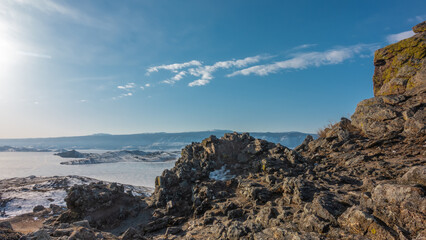 Fototapeta na wymiar Winter Siberian landscape. In the foreground is a picturesque granite rock with cracked slopes. In the distance - a frozen lake, a mountain range. Blue sky with clouds. Baikal