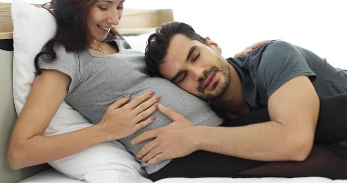 Tender loving husband listening to tummy of happy pregnant wife sitting on bed at home