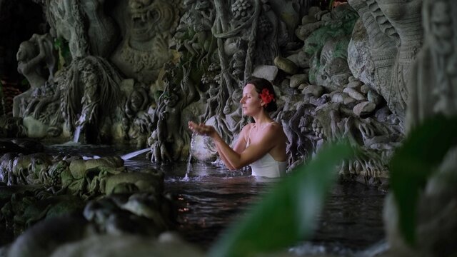 Beautiful woman with red flower in hair meditating and bathing in Bali water pond with Indonesian sculptures in tropics.