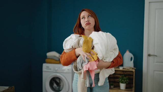 Young redhead woman unhappy holding dirty clothes at laundry room