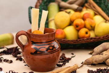 Jarrito de ponche de frutas navideño con caña y tejocote. canasta con frutas manzana, guayaba, canela y tamarindo. Sobre la mesa hojas de jamaica