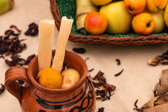 Jarrito de ponche de frutas navide&ntilde;o con ca&ntilde;a, tejocote y guayaba. Canasta con frutas, manzana. Sobre la mesa tamarindo y hojas de jamaica