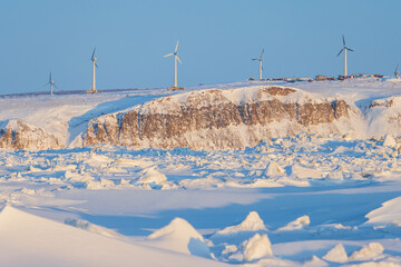 Wind farm in the Arctic on the sea coast. Wind turbines on a high rocky shore. Snow-covered ice. Green renewable energy in the Arctic. Modern electric power industry. Chukotka, Far North of Russia.