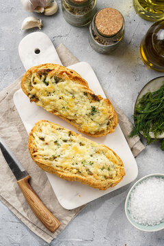 Two halves of garlic and butter bread - baguette on a marble board, sea salt, pepper, dill and garlic cloves on a grey concrete background, top view