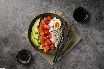 Poke bowl - green bowl with white rice, smoked salmon, cherry tomatoes and avocado, sesame seeds, soy sauce and chopsticks on a grey concrete surface, top view