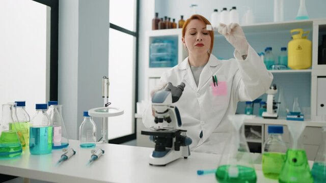 Young redhead woman wearing scientist uniform using microscope working at laboratory