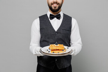 Young happy barista male waiter butler man wear white shirt vest elegant uniform work at cafe hold cheesecake waffles isolated on plain grey background studio portrait. Restaurant employee concept