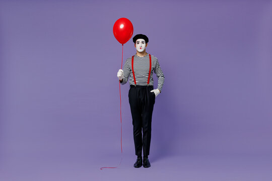 Full size body length vivid young mime man with white face mask wears striped shirt beret holding colorful air inflated helium balloon isolated on plain pastel light violet background studio portrait.