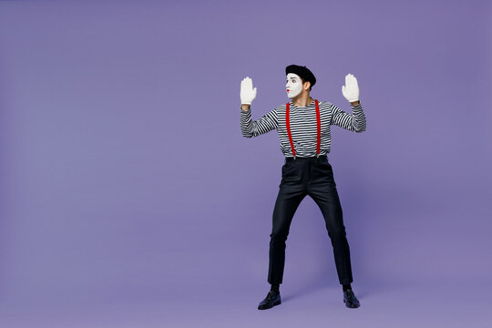 Full size body length young mime man with white face mask wear striped shirt beret look aside hands raised like touch invisible wall isolated on plain pastel light violet background studio portrait.