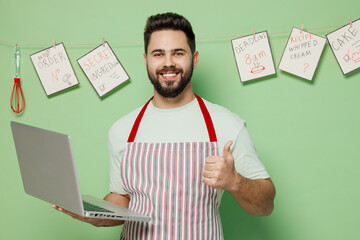 Young male chef confectioner baker man in striped apron hold use work on laptop pc computer show thumb up gesture isolated on plain pastel light green background studio portrait. Cooking food concept