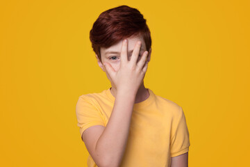 Portrait of a kid boy wearing casual t-shirt standing over isolated yellow background covering part of face with hand. Gestures. Childhood