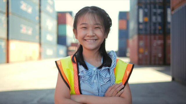 Portrait of Asian kid girl wearing reflective shirts smiling look to the camera at container cargo site. Concept of childhood development.