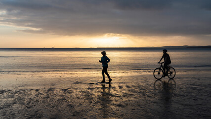 Silhouette image of two people running and cycling on a sandy beach at sunrise