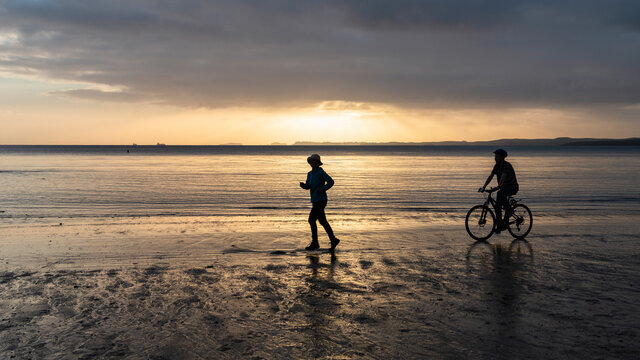 Silhouette image of two people running and cycling on a sandy beach at sunrise