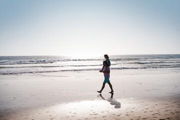 Handsome man father carrying young boy son. Happy dad holding child. Father and son walking on summer beach. Childhood and parenting concept.