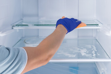 the hand of a cleaning service worker in a protective glove, wipes the glass shelves of the refrigerator with a sponge with cleaning foam