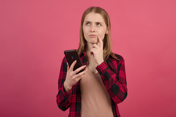 A young and attractive blonde Caucasian girl in a shirt using a mobile phone with a pensive look on a pink studio background.