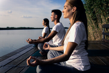 Group of people doing yoga exercises by the lake at sunset. 