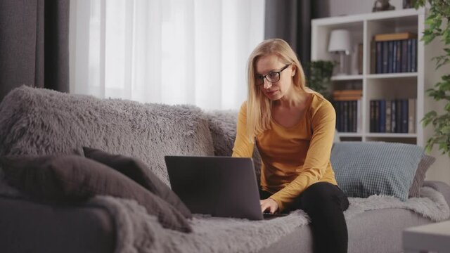 Pleasant caucasian woman in eyewear and casual clothes sitting on comfy couch and typing on wireless laptop. Modern technology during leisure time at home.