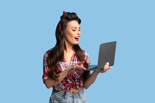 Sexy young pinup woman in plaid retro shirt using laptop over blue studio background