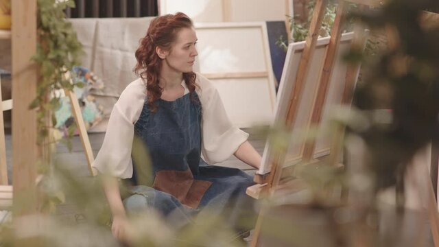 Full shot of young red-haired Caucasian woman wearing apron, sitting on floor and painting on canvas in modern art studio at daytime