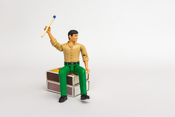 Miniature toy businessman figure sitting on a matchbox holding mach stick, on white gray background.