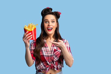 Excited young pinup woman in retro outfit holding package with yummy french fries on blue studio...