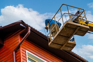A work carpenter repairs the roof of a wooden house at a construction site in a hydraulic lift of an aerial platform at a height.
