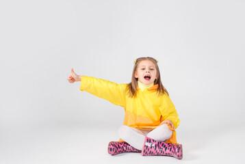 a little girl in a yellow raincoat and boots is sitting on a white background pointing her finger up. children's joy and happiness.