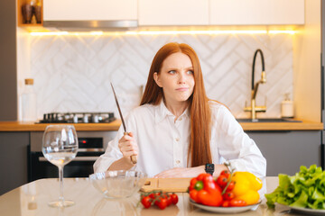 Obraz na płótnie Canvas Portrait of attractive redhead young woman with dissatisfied eyes holding big knife sitting at table with cutting board, looking away in light kitchen room with modern interior, close-up.