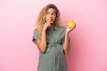 Girl with curly hair isolated on pink background pregnant and holding an apple while thinking