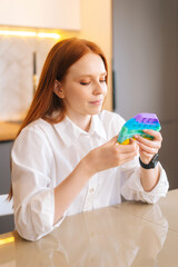 Vertical portrait of attractive redhead young woman playing fidget toy new trendy silicone toy anti stress pop it and simple dimple, sitting at table in kitchen room with modern light interior.