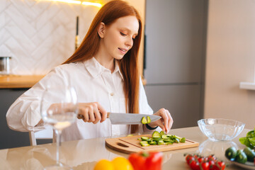 Obraz na płótnie Canvas Close-up side view of attractive young redhead woman cutting fresh cucumber cooking food salad sitting at table in kitchen room. Happy female cooking vegetarian dieting salad full of vitamins.
