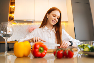 Portrait of cheerful young redhead woman cutting fresh tomato preparing food salad sitting at table in modern kitchen room. Happy female cooking vegetarian dieting salad full of vitamins.