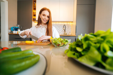 Portrait of young redhead woman cutting fresh yellow bell pepper preparing food salad sitting at table in modern kitchen room, looking at camera. Pretty female cooking dieting salad full of vitamins.