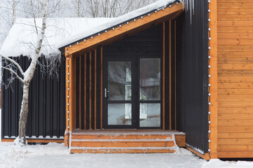 The house is in a modern style. Eco-friendly housing. Combination of wood and metal. Entrance door, panoramic windows. It's winter, there's snow and trees all around.